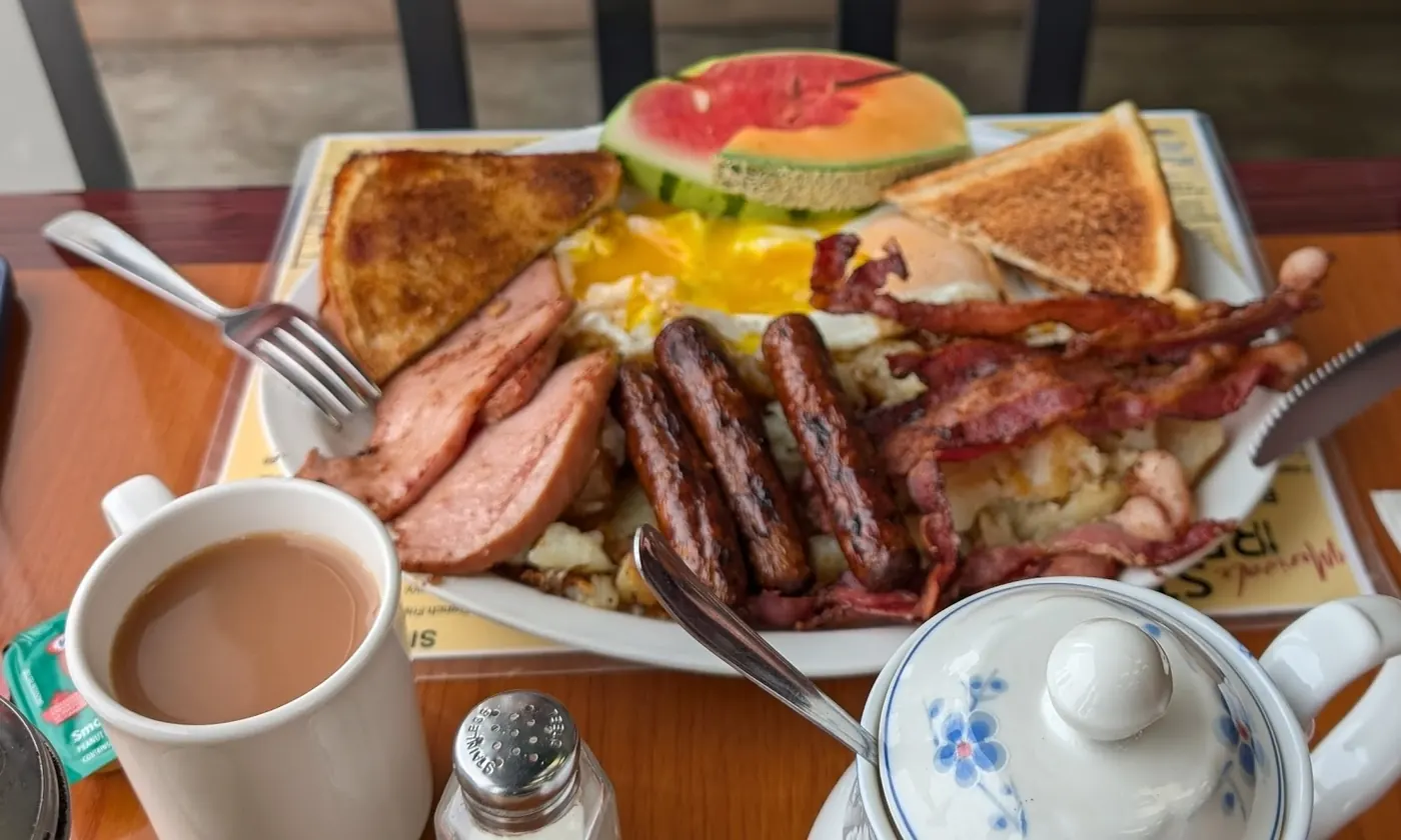 A big breakfast plate with ham, sausages, bacon, eggs and toast, with a mug of tea and a teapot beside it