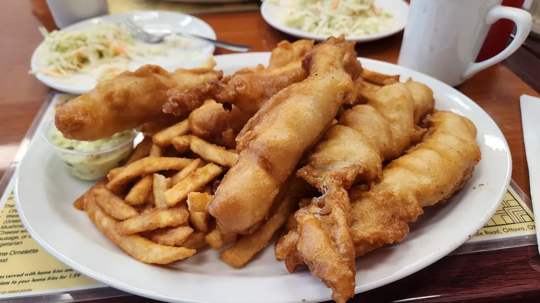 Close-up of a plate piled with battered fish pieces and fries, with tartar sauce and coleslaw behind